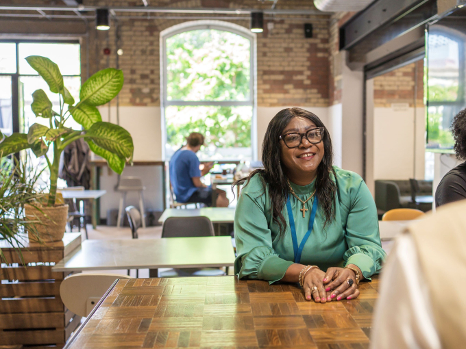 Professional in green top working at a wooden table in a bright co-working café with natural light, brick walls, and indoor plants — modern workspace with collaborative atmosphere.