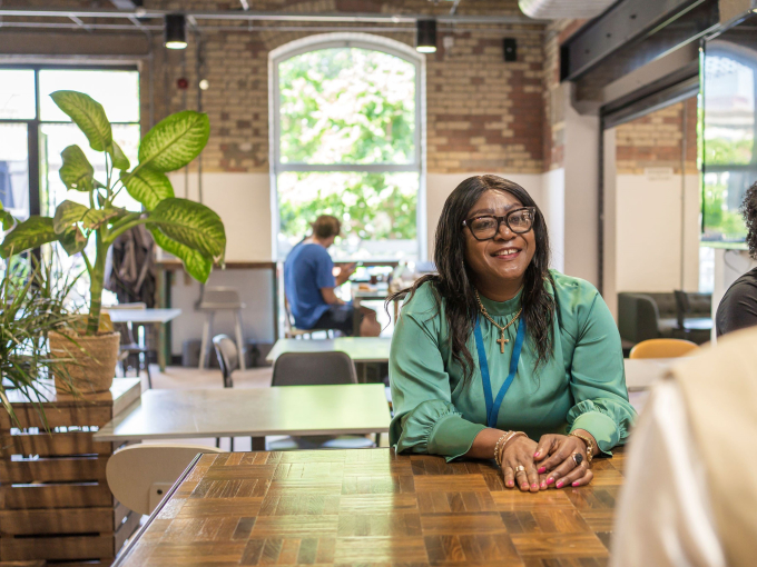 Professional in green top working at a wooden table in a bright co-working café with natural light, brick walls, and indoor plants — modern workspace with collaborative atmosphere.