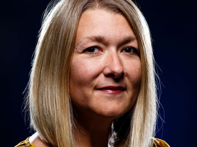 A smiling lady dressed in a patterned top, looking at the camera, against a black backdrop.