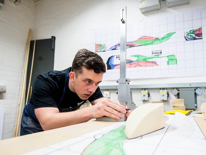 A young man stoops over a table, eyes fixed on a metal height gauge; the wall behind him displays plans for a sports car chassis.