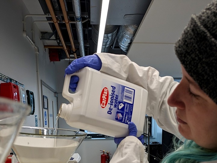 Wearing a woollen hat, a white labcoat and rubber gloves, Rebecca Macfarlane pours liquid from a tub labelled De-ionised Water into a plastic funnel.
