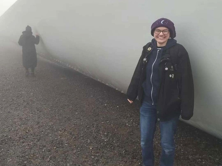 Wearing a purple hat with a crescent moon in its centre, Rowan Jack Moses stands smiling in front of a large, long shape that might be the hull of a ship.