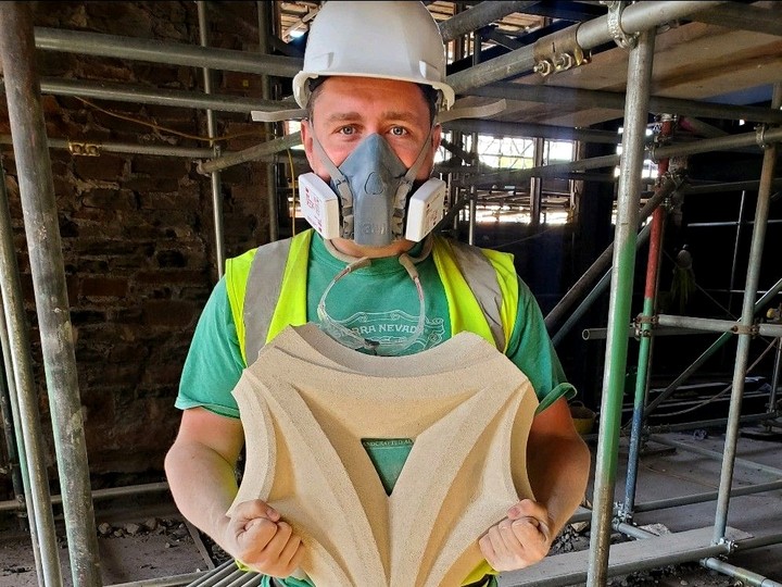 John Langstone in a hard hat and respirator face mask; standing in front of scaffolding, he holds up a piece of stone tracery with both hands.