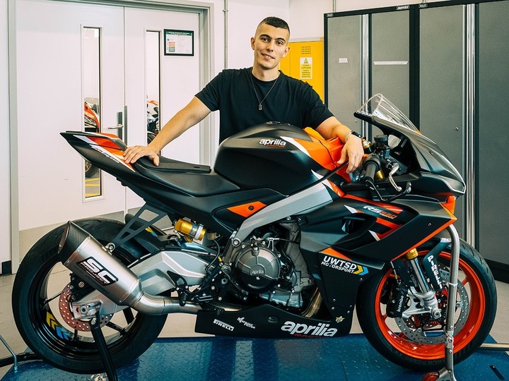 Vinicius Dreher standing and smiling, hands resting on the saddle and fuel tank of an Aprilia racing motorbike.
