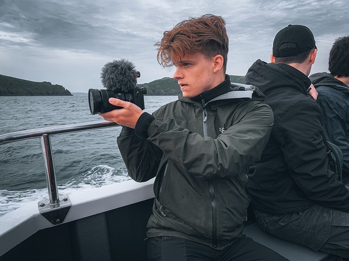 On a grey day, Joshua Knight sits near the stern of a boat and turns towards the sea, holding up a camera with a microphone attachment.