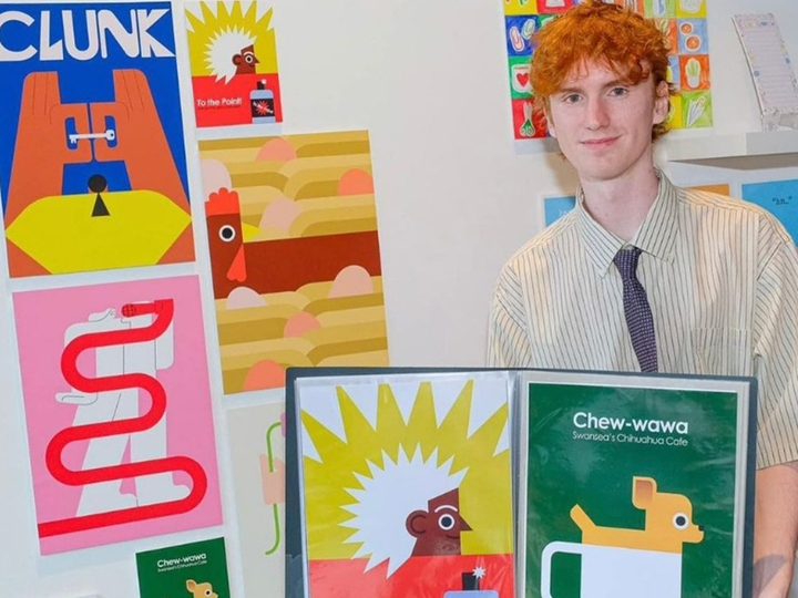 Evan Elias smiles slightly towards the camera as he stands by an exhibition of his illustrations.