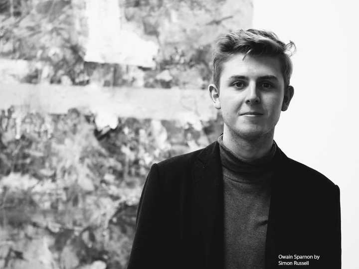 A black-and-white photo of Owain Sparnon wearing a black coat over a polo-neck; he is standing in front of an abstract, textured artwork.