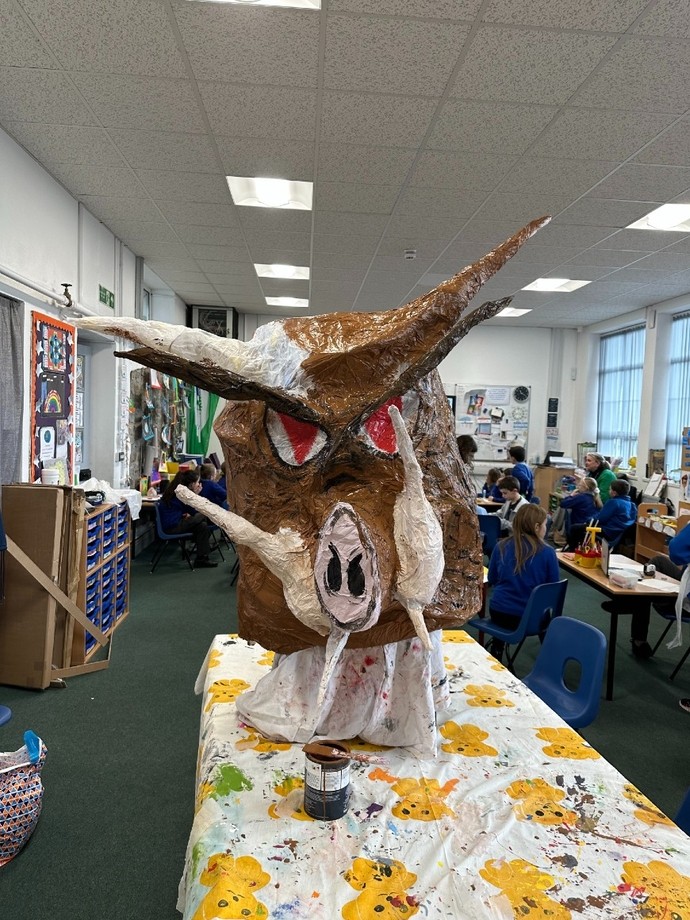  A puppet of a brown wild boar in the process of being painted with brown paint; schoolchildren are sitting in the background.