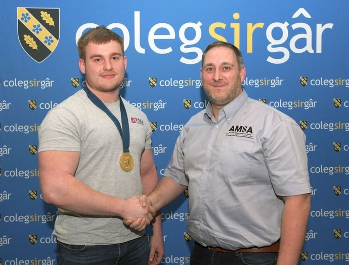 Gan wisgo medal aur gyda balchder, mae’r myfyriwr Kian Lloyd yn ysgwyd llaw â chynrychiolydd o AMSA (yr Academi Sgiliau Gweithgynhyrchu Uwch) wrth ddathlu ei fuddugoliaeth yn y gystadleuaeth.