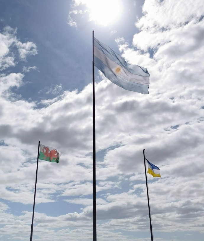 Tair baner wedi’u codi ar fastiau o flaen cefndir heulog.