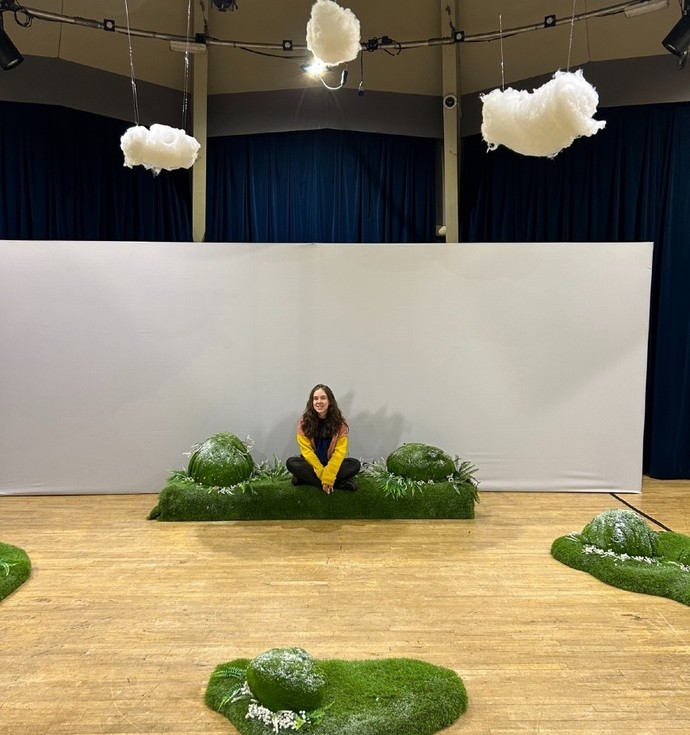 Smiling, Lleucu Williams sits cross-legged on a hump of fake grass on the stage set. 