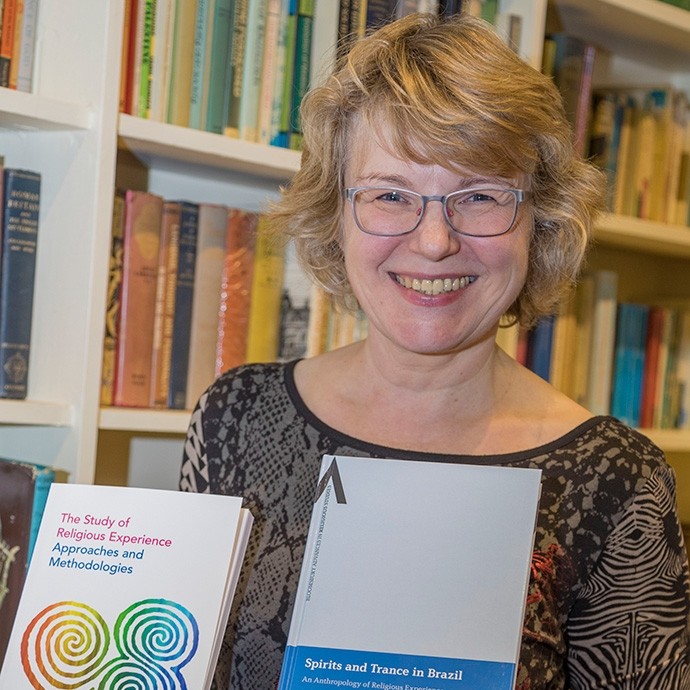 Yn sefyll o flaen cwpwrdd llyfrau, mae’r Athro Bettina Schmidt yn gwenu tuag at y camera wrth ddal dau o’u gweithiau ei hun: “Astudiaeth o Brofiad Crefyddol – Dulliau a Methodolegau”; hefyd, “Ysbrydion a Llesmair ym Mrasil”.