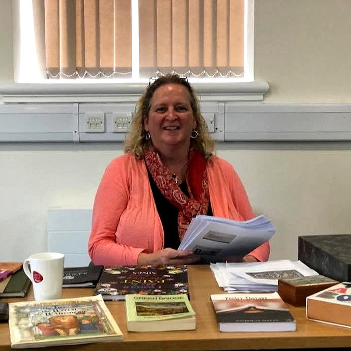 Dr Jessica Clapham yn eistedd wrth fwrdd gydag amrywiaeth o lyfrau a phapurau drosto.