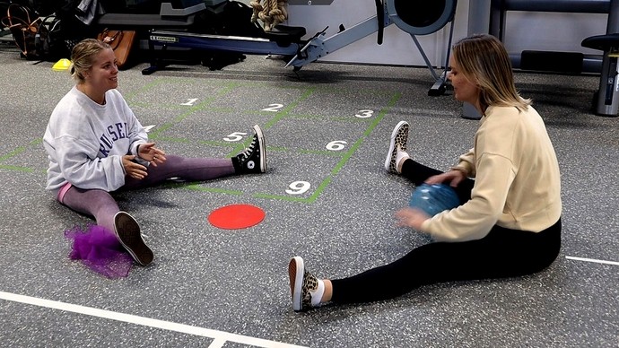 Two young women sit on the floor of a gym, rolling a ball between them. 