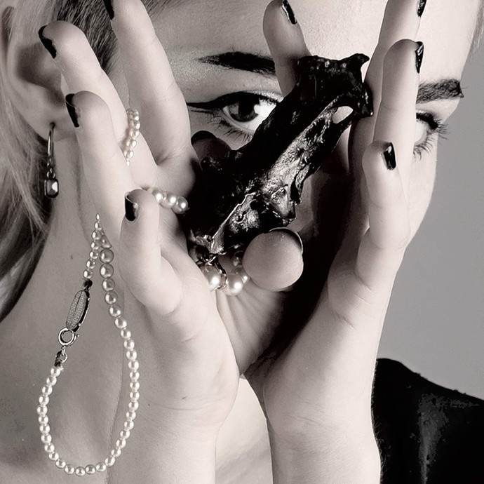 A young woman holds a dried black shark egg case and a string of white pearls towards the camera in a desaturated photograph; her eye looks towards the viewer over the edge of the egg case.