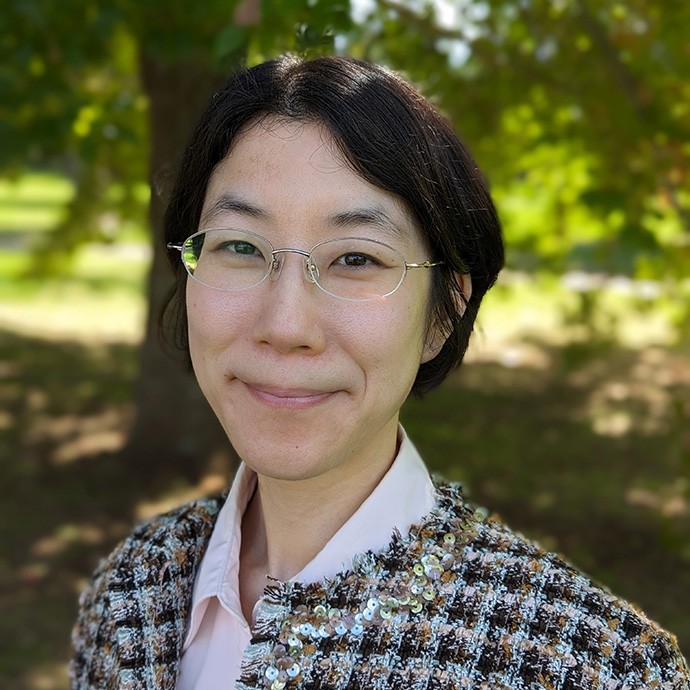 Mae Dr Katherine Ngo yn gwenu tuag at y camera ar ddiwrnod heulog gyda choeden ddeiliog tu ôl iddi.