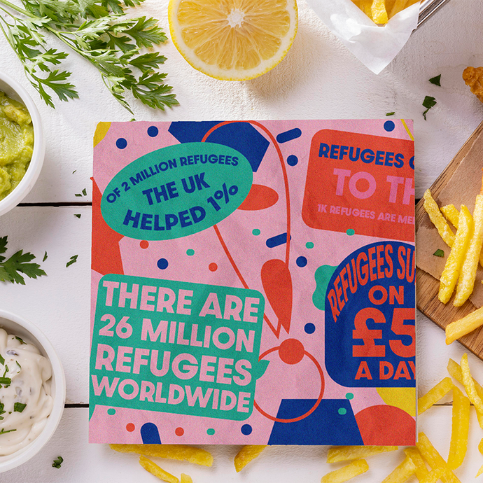 On a table spread with fish and chips, a lemon, and tartar sauce, a square card reads: of 2 million refugees the UK helped 1%; there are 26 million refugees worldwide; more text overlaps the side of the card, which uses a colour scheme of green, blue and red on a pink background.  