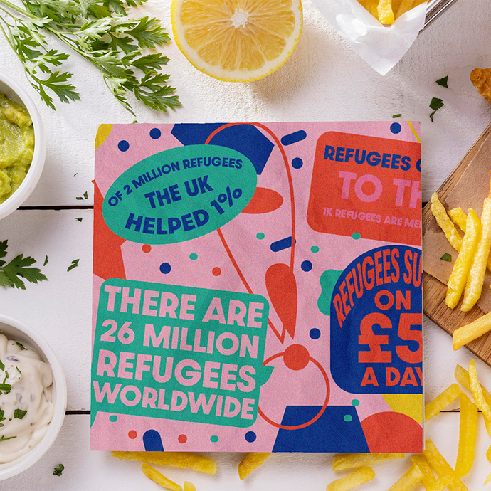 Ar fwrdd ac arno bysgod a sglodion, lemwn, a saws Tartar, mae cerdyn sgwâr yn dweud: 2 million refugees the UK helped 1%; there are 26 million refugees worldwide; mae mwy o destun yn gorgyffwrdd ochr y cerdyn, sy’n defnyddio cynllun lliw o wyrdd, glas a choch ar gefndir pinc.  