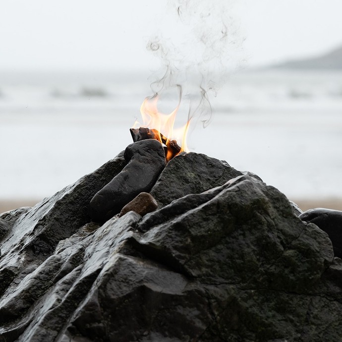 Fflamau’n codi o begwn riff creigiog ar draeth; mae ongl agos y llun yn gwneud i’r riff edrych fel llosgfynydd.