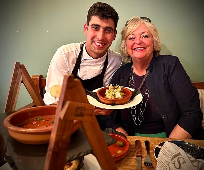 A smiling man in a chef outfit next to a happy, smiling lady with plates of food in front of them.