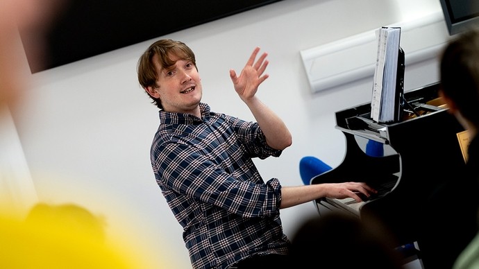 Saif darlithydd gydag un llaw ar allweddell piano, a’r llall wedi’i godi mewn ystum sy’n dangos pwyslais wrth iddo esbonio rhywbeth i’w gynulleidfa o fyfyrwyr.