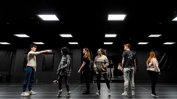 Nine students in black stand in a dark room with a single white spotlight; they raise one arm in the air and look solemn.