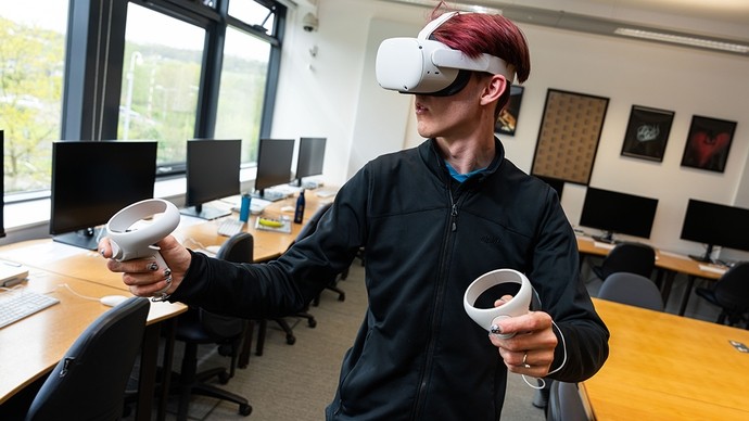 Mae myfyriwr yn sefyll o flaen dosbarth yn gwisgo penset Oculus Quest ac yn gafael dau reolydd.
