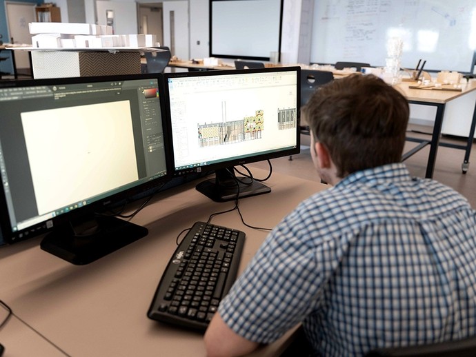 Eistedda myfyriwr o flaen monitorau deuol; mae un monitor yn dangos Adobe Photoshop a’r llall yn dangos cynllun adeilad.