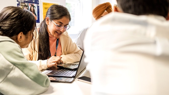 Young people sit around a table; one student checks her phone as it rests on an open laptop.