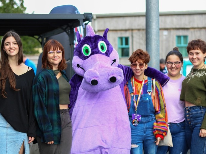 Mae person mewn gwisg draig borffor fwythus yn sefyll mewn llinell o fyfyrwyr gyda’i freichiau o gwmpas y ddau sy’n agosach ato; mae’r fyfyrwraig ar ei chwith yn gwisgo cortyn Pride a siwmper enfys.