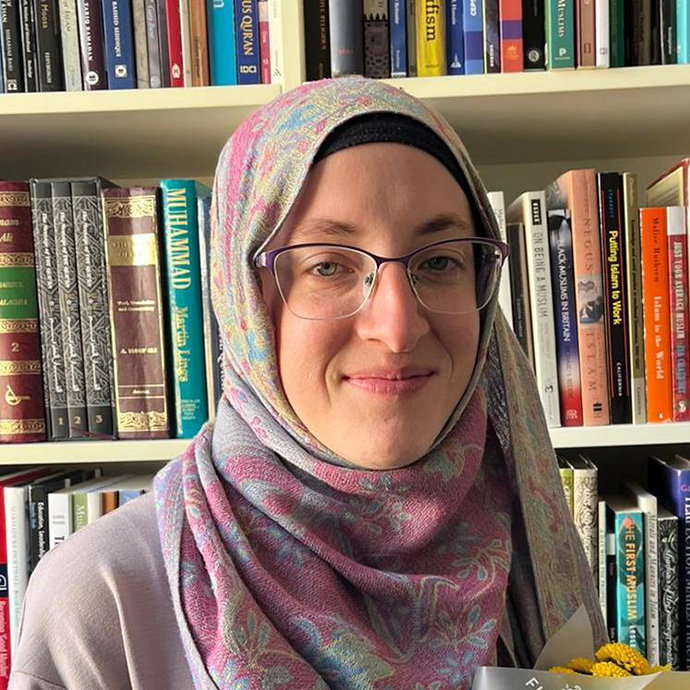 Yn gwisgo hijab a sbectol binc golau, mae Dr Laura Jones yn edrych tuag at y camera ac yn gwenu; mae'r llyfrau ar y silffoedd y tu ôl iddi yn cynnwys teitlau ar ddiwinyddiaeth Islamaidd. 
