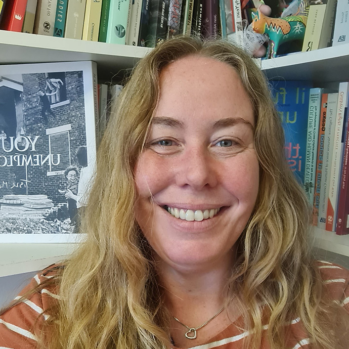 Yn sefyll o flaen silffoedd llyfrau llawn, gyda gwallt melyn hir ac yn gwisgo loced arian siâp calon, mae Dr Jessica Pitman yn gwenu i mewn i'r camera.  