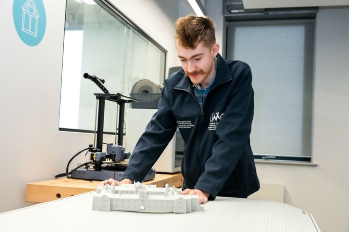 A man in a branded jacket looking down at a white plastic model of a building in a lecture room.