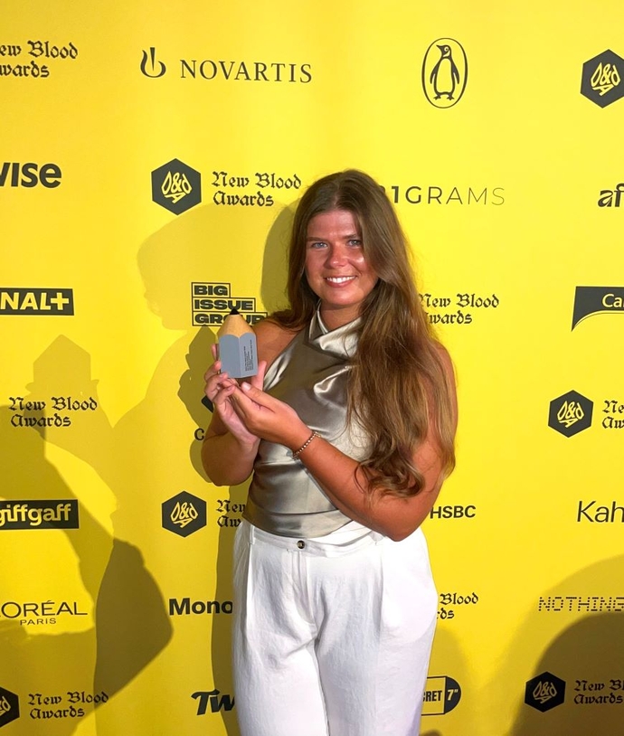 A happy, smiling student standing against a bright yellow branded background, holding her award.