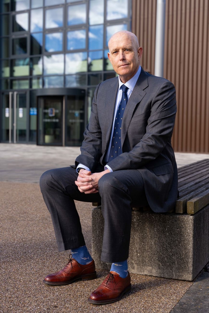 A man seated on a stone bench, with his arms resting on his knees, in front of a red brick and glass building.