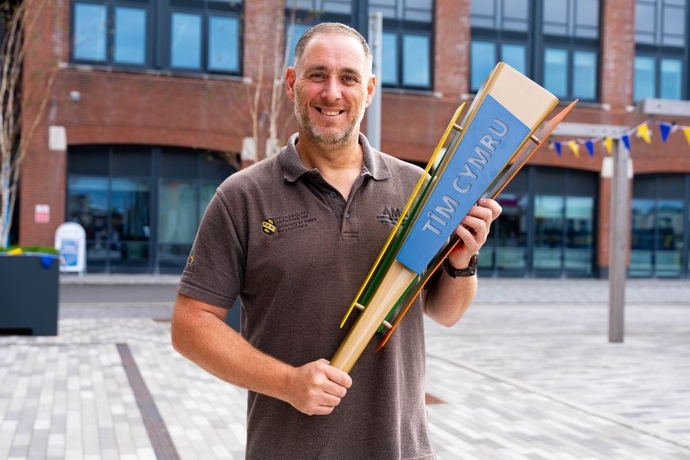 A man in a brown T-Shirt holding a wooden replica torch, standing in front of a red brick building.
