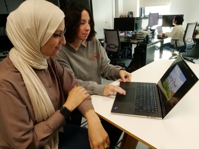 Two University staff sat at a white desk looking at technology.