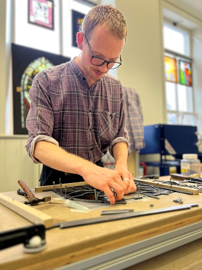 An artist working on stained glass with more examples on the walls behind him.