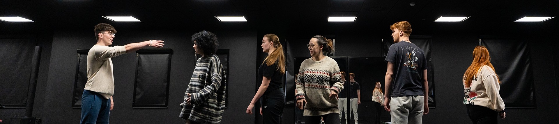 Six students assume different attitudes under the lights of a black-walled rehearsal room.