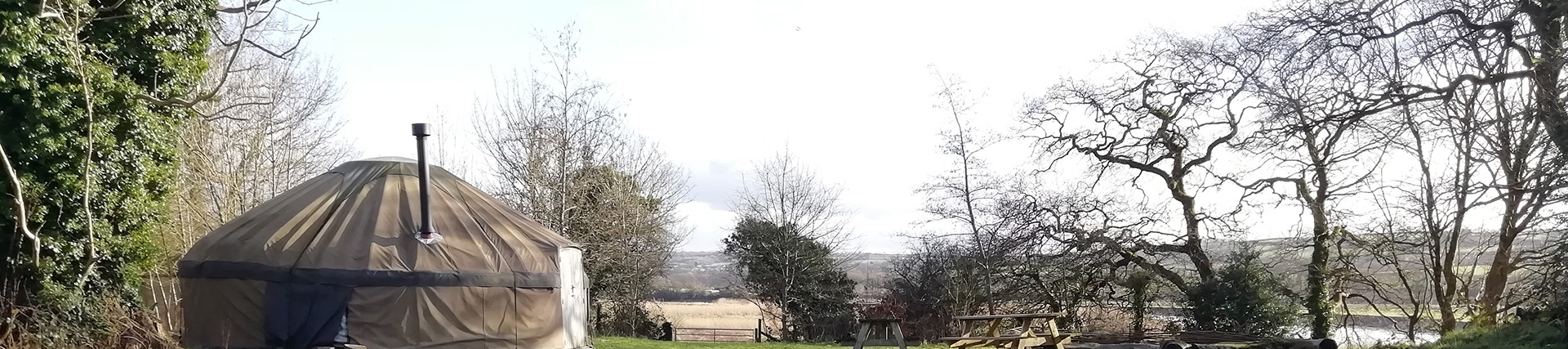 A large canvas yurt in a small field; the yurt has a metal chimney; there is a seating area outside with logs and picnic benches; river meadows and the Towy itself are visible in the background. 