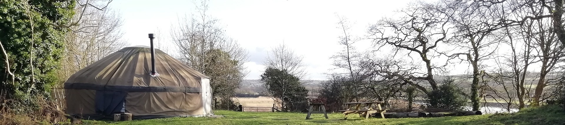 A large canvas yurt in a small field; the yurt has a metal chimney; there is a seating area outside with logs and picnic benches; river meadows and the Towy itself are visible in the background. 