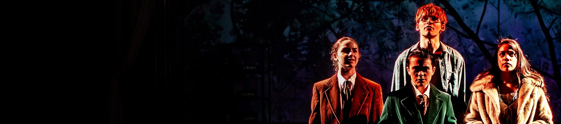 Four young actors stand in shadowy light; the stage curtain is to the left and behind them the scenery includes the black shapes of trees against a nightime backdrop.