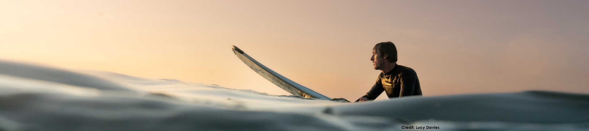 A surfer stands in the sea waiting for a wave against a pale yellow sky; photo credit Lucy Davies.
