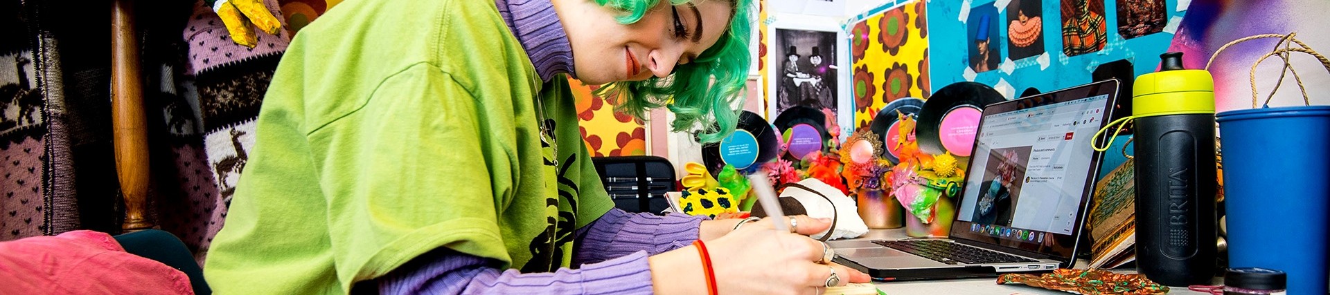 A student in bright clothes with vivid green hair smiles slightly as she makes notes at a table in a  colourful art studio. 