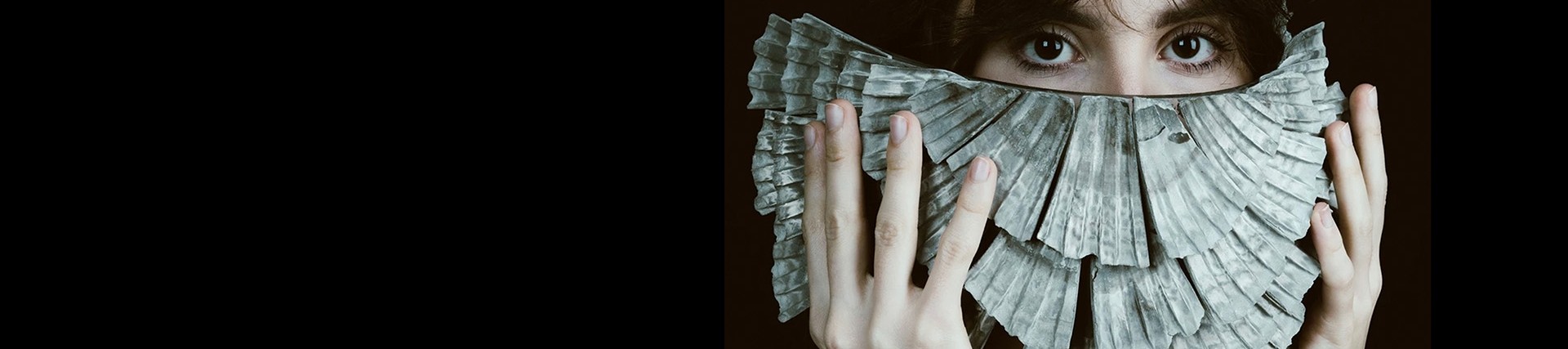 A young woman holds a half-circle of layered shells over the lower half of her face; her eyes look towards you.