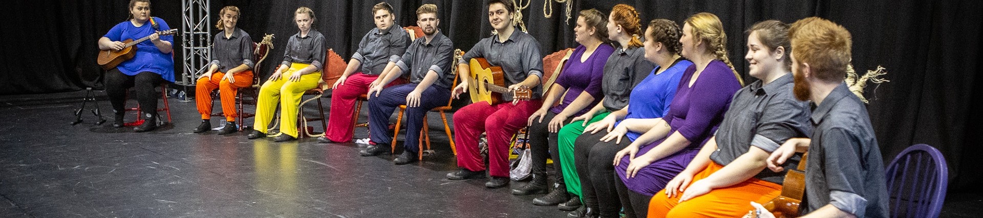 A long line of seated young people, a few with acoustic guitars, all wearing grey shirts and brightly coloured trousers.
