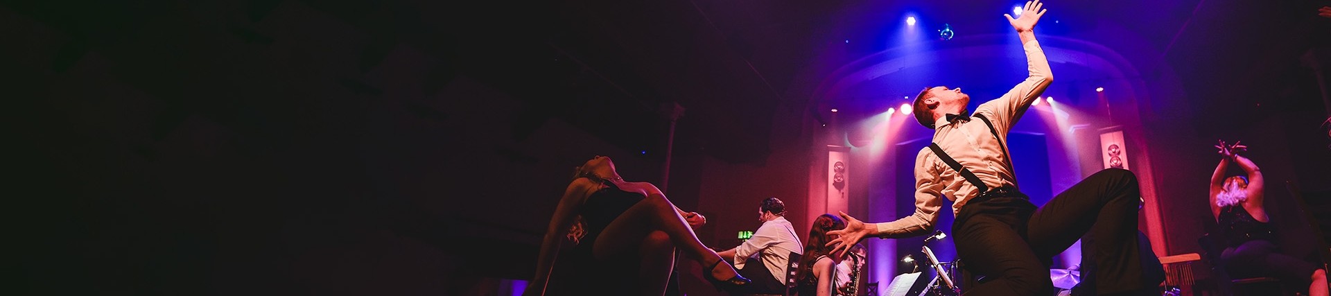 A nightclub scene illuminated by purple backlights; a young actor in a bow tie and black braces over a white shirt is captured in an exultant dance pose.