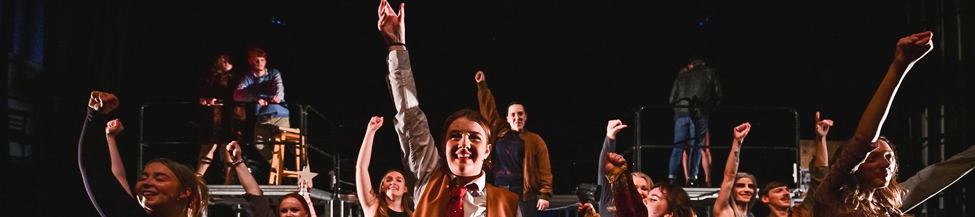 A young cast of twelve people smile towards the camera and raise their arms; some are cross-dressed; a couple stand on a gantry at the back and kiss. 