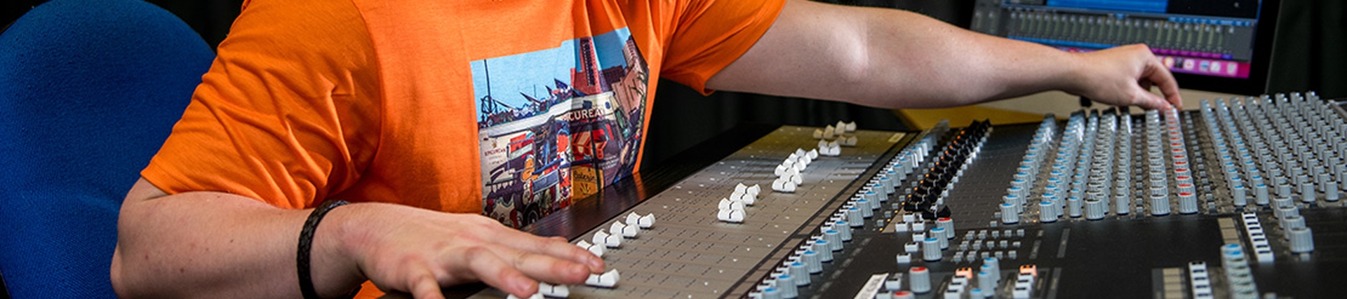 A young man in an orange t-shirt and baseball cap smiles as he makes adjustments to the dials and sliders on a mixer board.