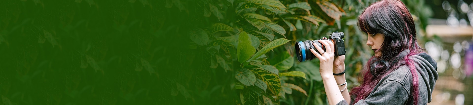 A young woman holds a camera looking in the direction of some tropical plants with large green leaves.